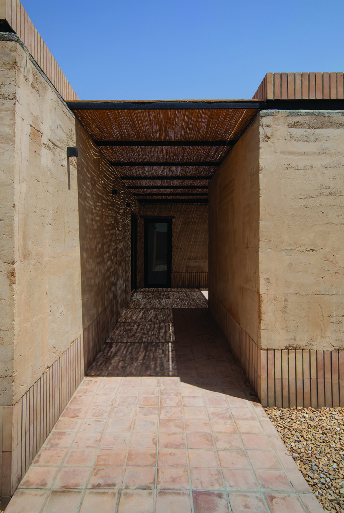 View through a narrow covered passageway with rammed earth walls on both sides and a reed canopy overhead, with dappled light on brick flooring