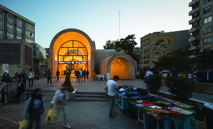 Dusk view of the plaza entrance with warm lighting illuminating the arched canopy, pedestrians with shopping bags, and vendors displaying clothing on tables