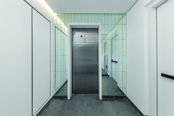 Elevator lobby with frosted glass tile walls, stainless steel elevator doors, and grey stone floor