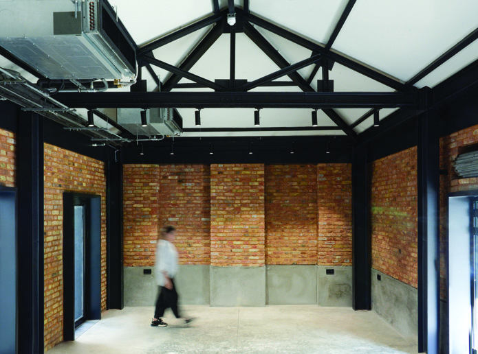 Third-floor event space with exposed brick walls, steel trusses supporting a translucent ceiling, concrete base, and a person walking through