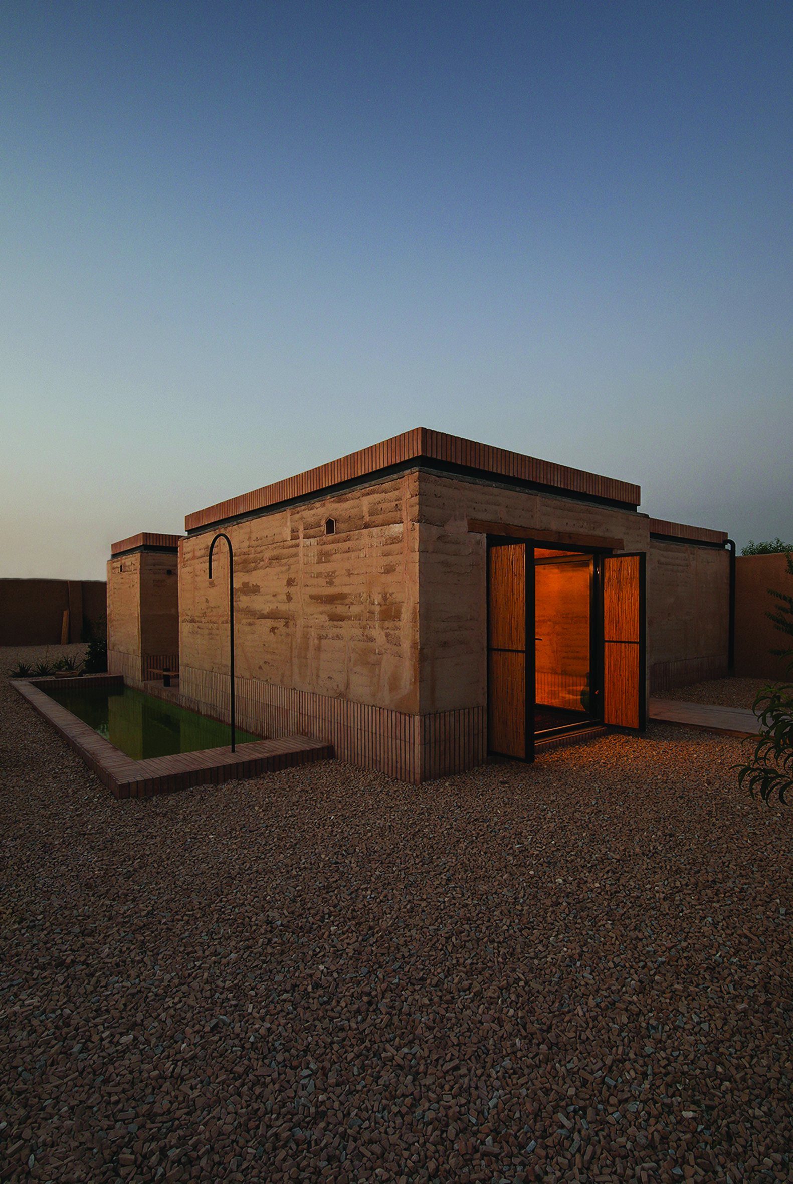 Evening view of rammed earth facade with a green-lit water pool reflecting the building, and warm light emanating from the interior