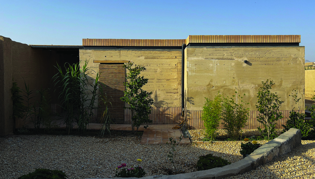 Daytime view of the house from the garden showing rammed earth walls with horizontal layers visible, reed door screen, and young plantings in the gravel yard