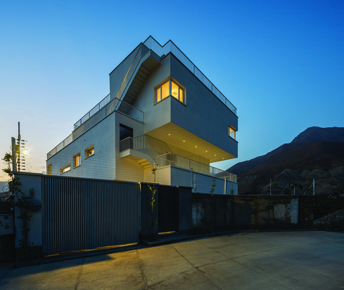 The Saman building at twilight from the north, showing the illuminated staircase and windows against the blue mountain backdrop, with the distinctive angular massing visible