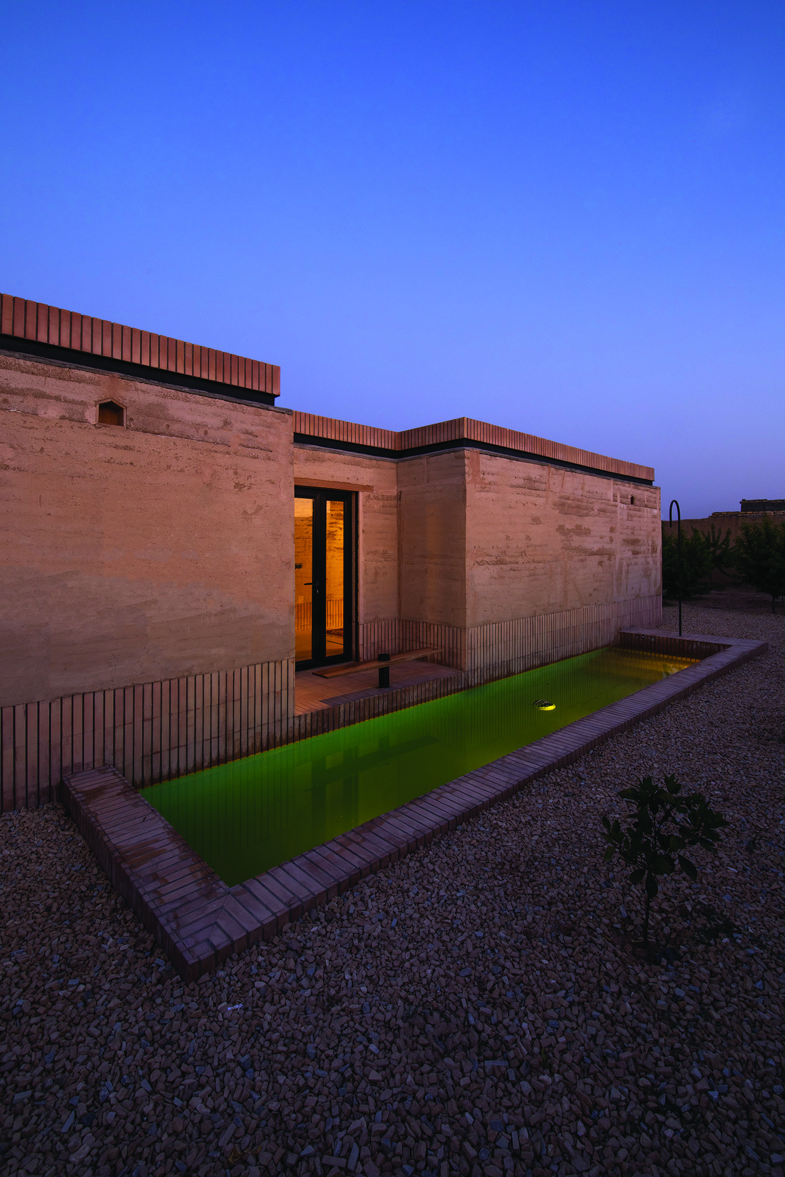 Twilight view of the south facade showing the rammed earth house with a green-lit pool, warm interior light, and clear sky