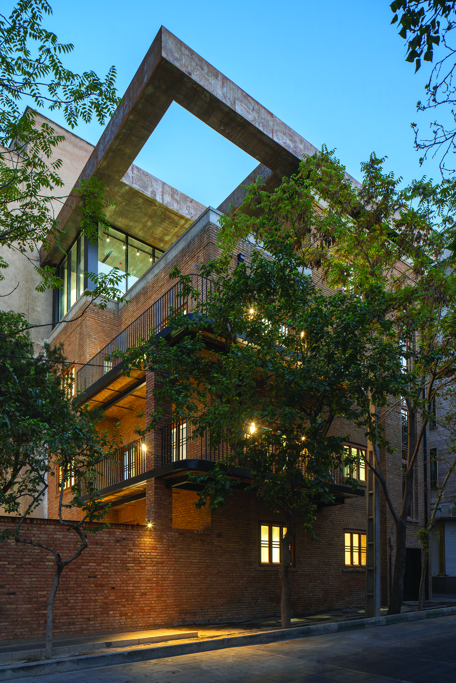 Close-up of the facade showing the bold cantilevered concrete frame extension above the original brick building, with illuminated balcony and trees