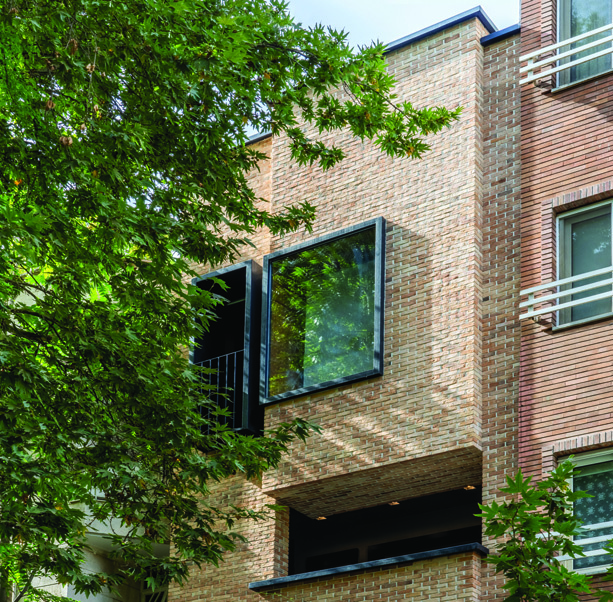Close-up of the upper facade showing a protruding glass window bay framed in dark metal against the warm brick wall, with tree foliage in foreground
