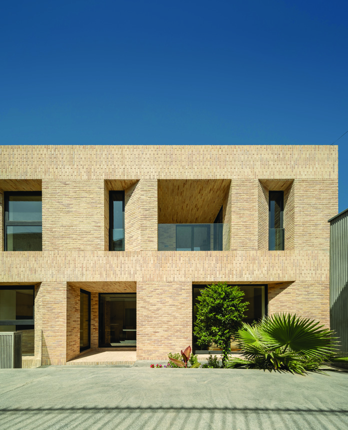 Street-facing facade of House No. 10 Jolfa showing a two-story brick building with recessed windows, terraces, a courtyard tree, and palm plant visible at ground level