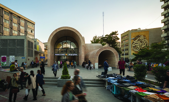 Frontal view of the plaza at dusk showing the main arch with glass wall glowing warmly, the metro entrance signage reading Meydan-e Jahad, pedestrians, and street vendors in the foreground