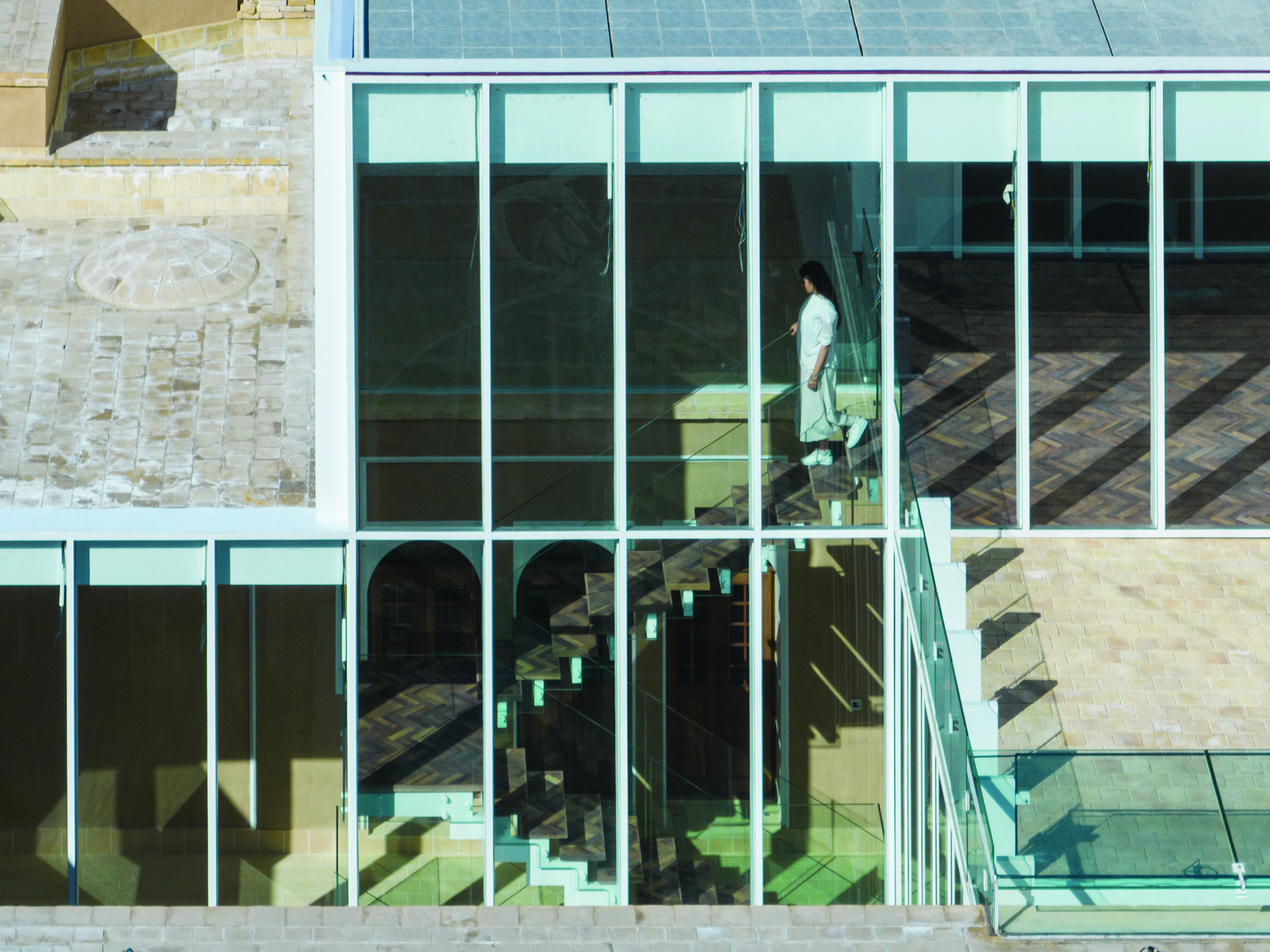 Close-up view of the glass facade showing a woman ascending the floating staircase, with the old courtyard reflected in the glazing