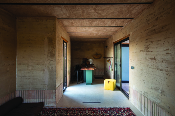 Interior hallway connecting to the kitchen area with rammed earth walls, colorful furniture, and natural light from glass doors on both sides