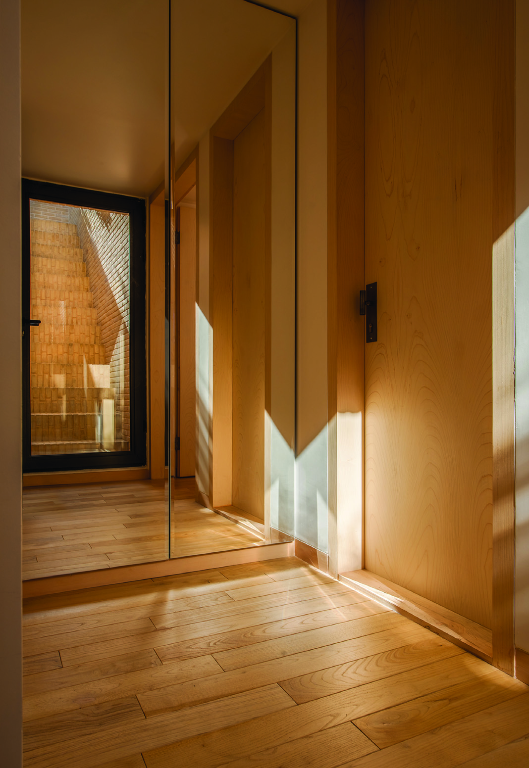 Hallway with wooden floors and paneling, a mirror reflecting the brick-clad stairwell, and dramatic diagonal sunlight patterns