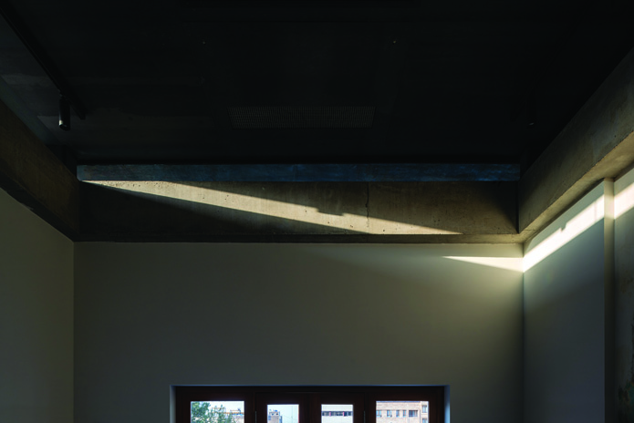 Interior room with a dramatic beam of natural light cutting across the concrete ceiling and wall, with windows visible in the background