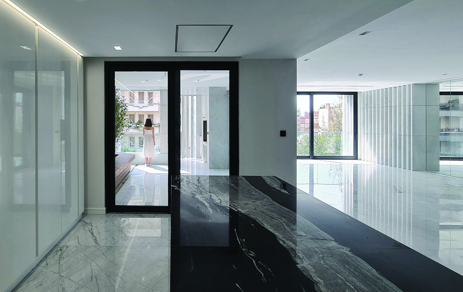 Interior of a Sayeh Building apartment showing a spacious living area with black and white marble flooring, glass doors opening onto a terrace where a woman stands, and stone louvers visible through the window