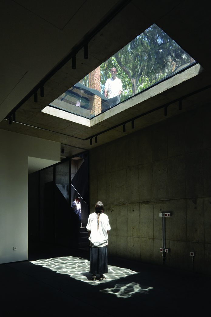 Interior space with exposed concrete walls, a large glass skylight overhead, a woman standing beneath it gazing up, and dappled water-like light patterns on the floor