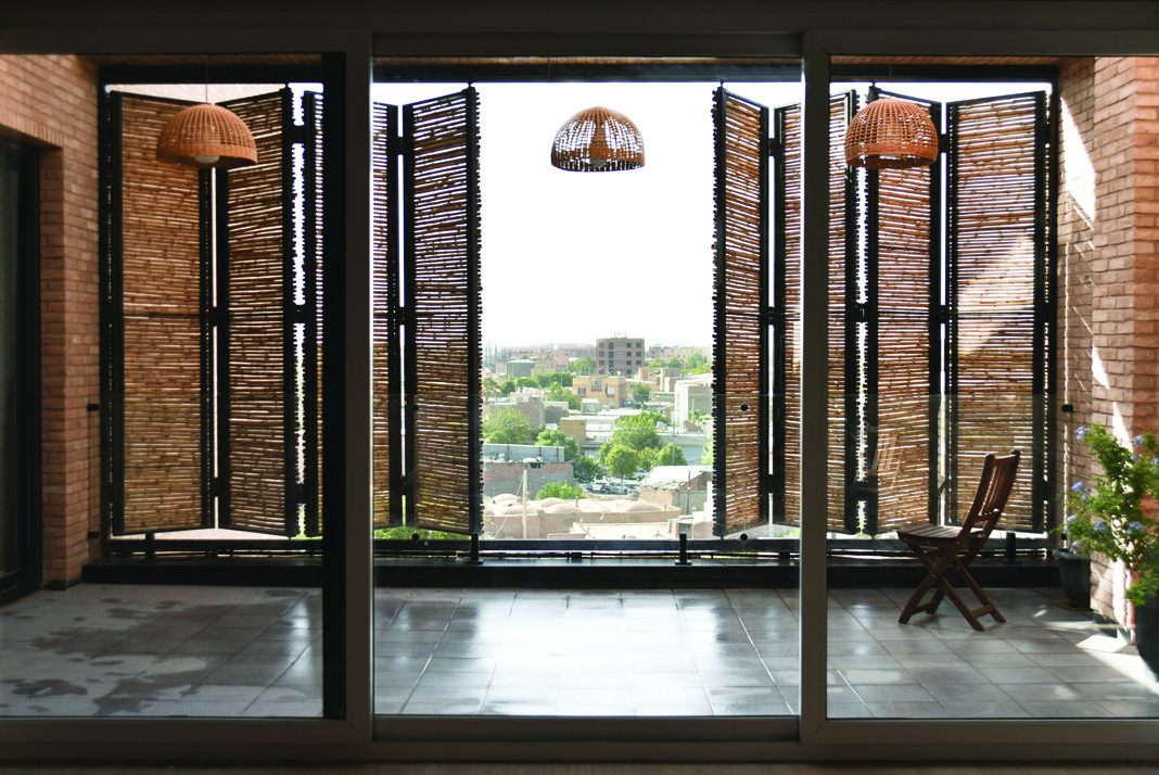 Interior view looking out through floor-to-ceiling glass doors onto the terrace, with open reed panels and wicker pendant lamps, the Kerman cityscape visible beyond