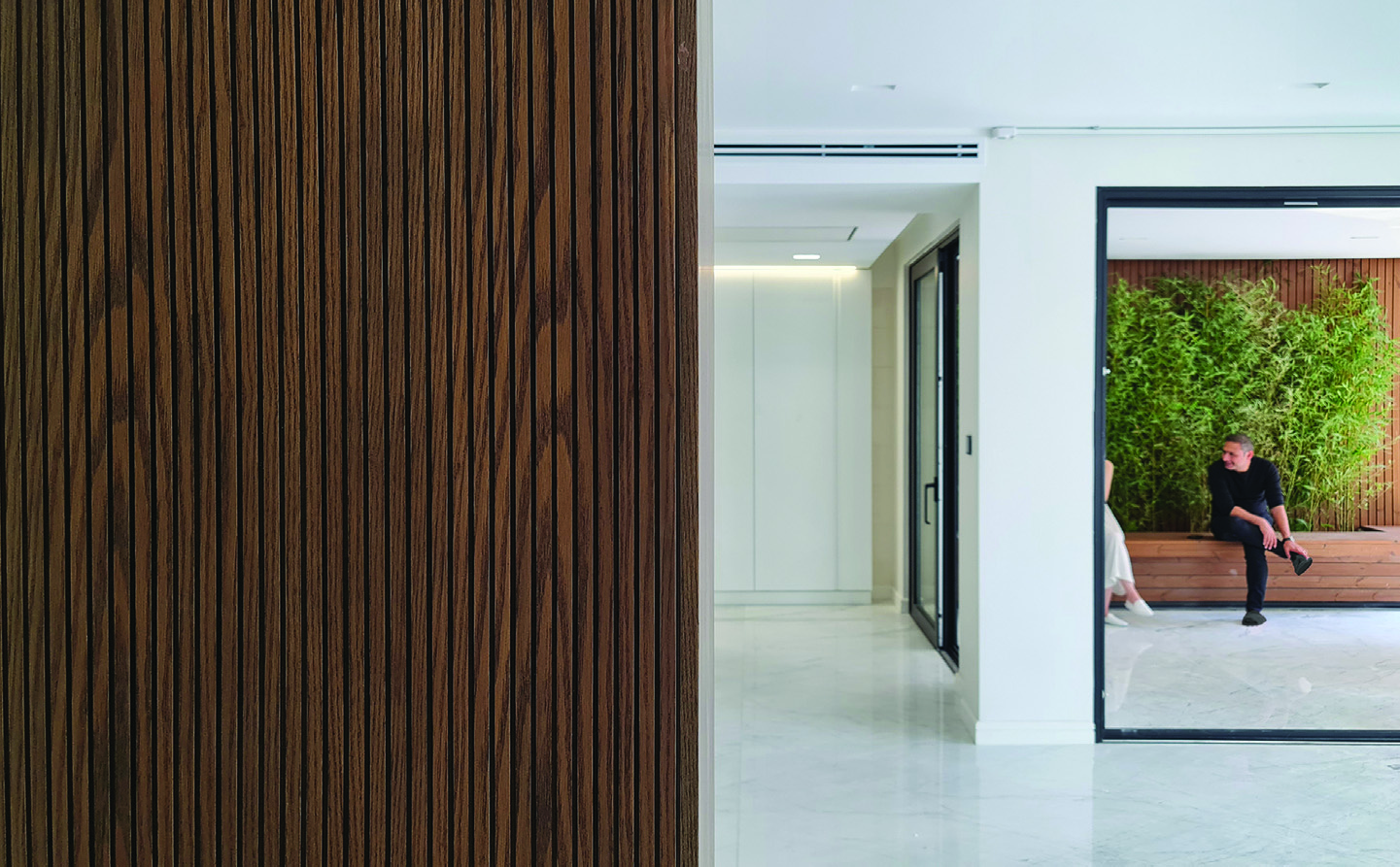 Interior view past a dark wood-paneled wall toward a glass door opening onto a green courtyard, where a man sits among bamboo plants