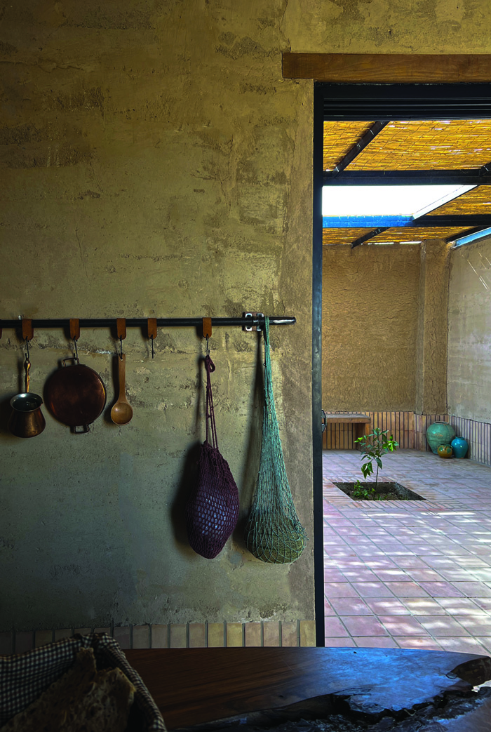 Kitchen detail showing hanging copper pots, woven bags, and rammed earth walls, with a view through to the reed-canopied courtyard beyond