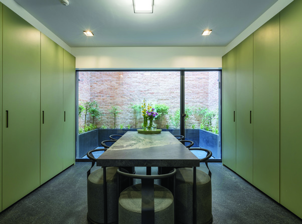 The kitchen-dining room with sage green seamless cabinet doors, a dark marble dining table, and a large window opening to the planted courtyard with brick walls