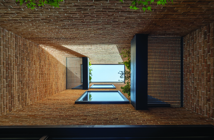 View looking up through the brick-clad lightwell showing a vaulted brick ceiling, glass floor panels, and wooden privacy screens admitting dappled light