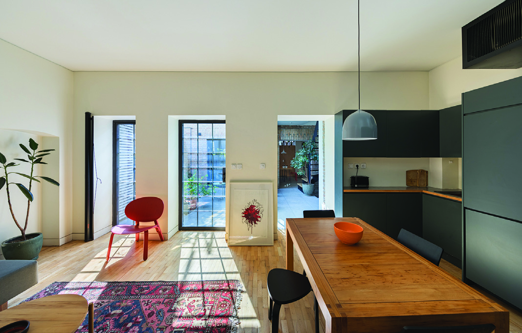 Open-plan living room and kitchen with wooden floors, a Persian rug, red accent chair, and three tall windows looking onto the courtyard