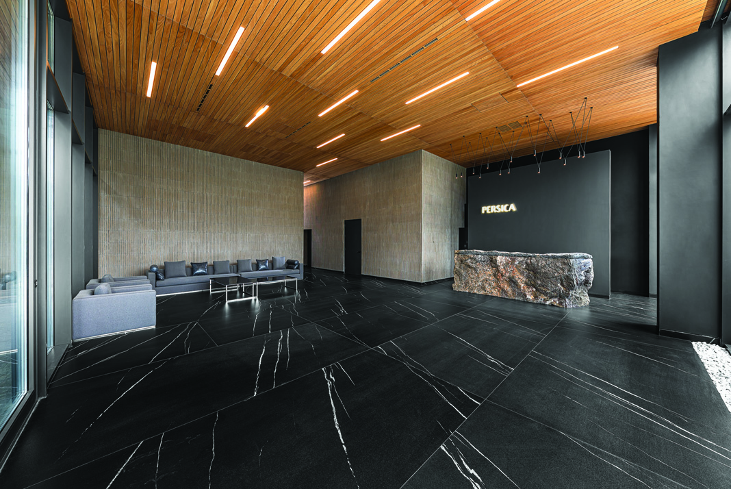The Persica lobby with a raw stone reception desk, 'PERSICA' illuminated sign, black marble floor, and warm wooden slatted ceiling
