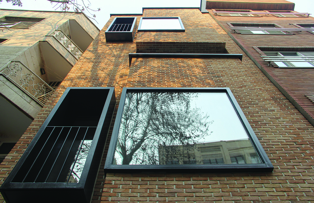 Low-angle view looking up at the brick facade showing asymmetric window openings, a protruding glass bay, and metal balcony railings against an overcast sky