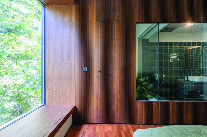 The master bedroom featuring warm wood slat wall paneling, a large window overlooking tree canopy, and a glass partition revealing the green-tiled bathroom beyond