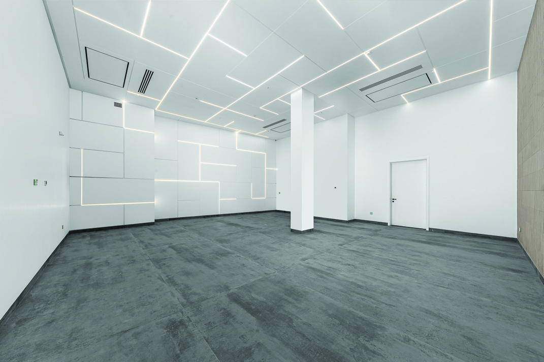 Multi-purpose hall with geometric LED ceiling lighting, white acoustic wall panels, and grey concrete floor