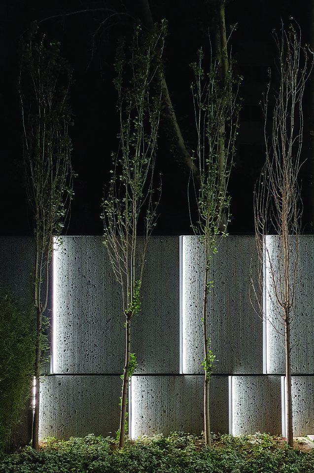 Night view of the building's garden wall with uplighting illuminating young trees against textured stone panels and low ground cover