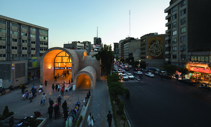 Night view of the lit plaza from across the street showing the glowing arched structure, traffic, and the urban context of surrounding buildings