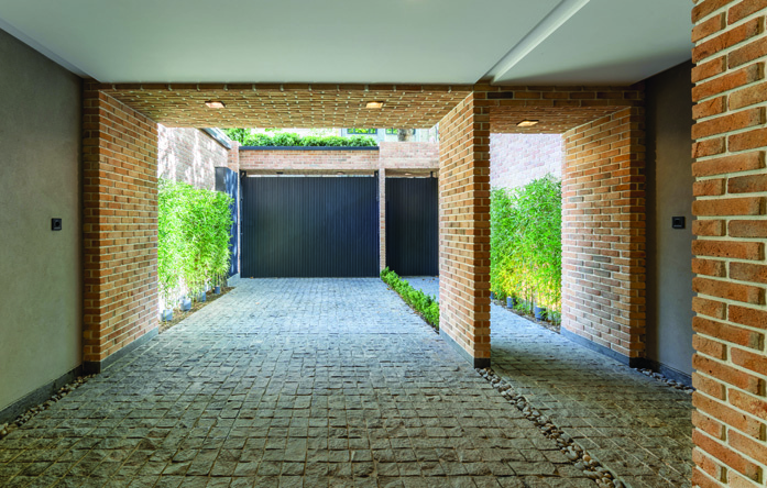 The ground-level parking corridor with brick column piers, cobblestone floor, greenery on both sides, and the metal gate at the far end