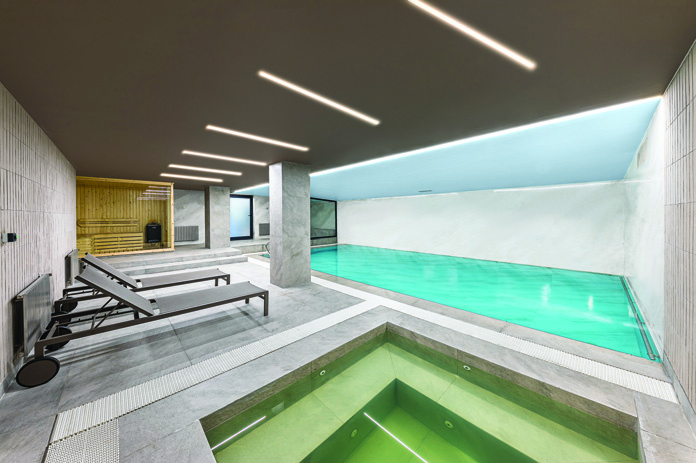 Indoor swimming pool and jacuzzi in the basement level with recessed linear lighting and sauna visible in the background