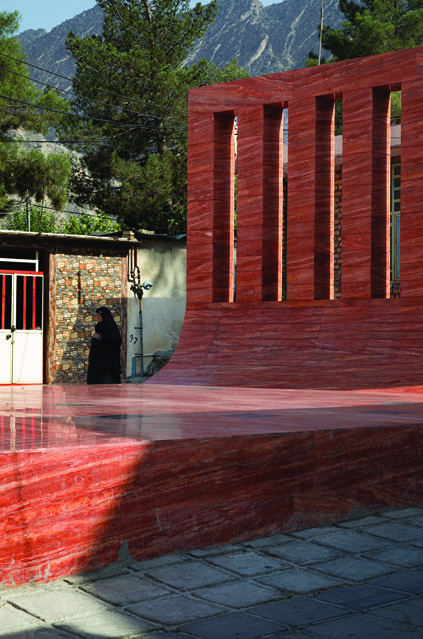 View of the vertical red stone wall with eight tall openings revealing cypress trees behind, the curved transition from wall to ground plane visible, mountains in the background
