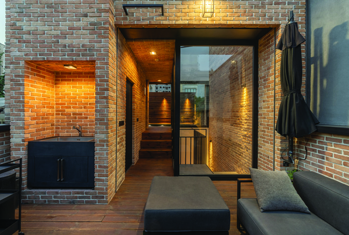 The roof terrace at dusk with warm lighting, a built-in BBQ area recessed into brick wall, outdoor sofa, and a glass door leading to the stairwell