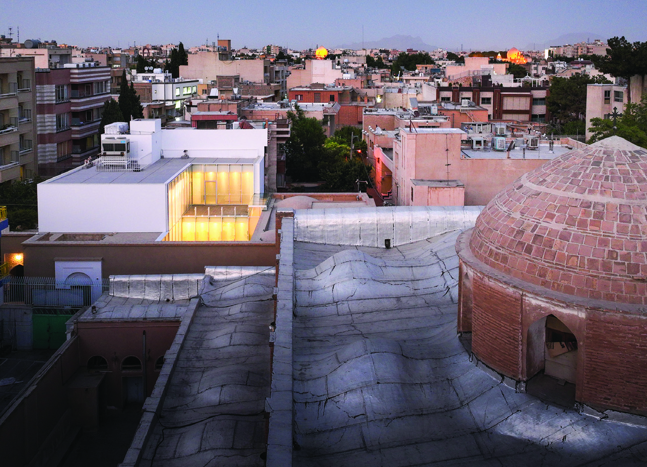 Twilight view from above showing the illuminated glass pavilion of Once Again Home with the brick dome of the neighboring church in the foreground