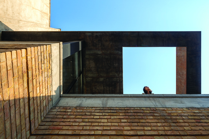 Rooftop terrace with warm brick surfaces and a bold concrete frame opening to the sky, a person gazing upward in the distance