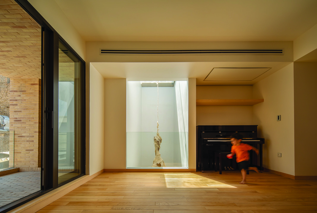 A room with hardwood floors, a piano in the corner, a child in motion, and a large glass wall looking into the brick-lined lightwell with a hanging sculpture