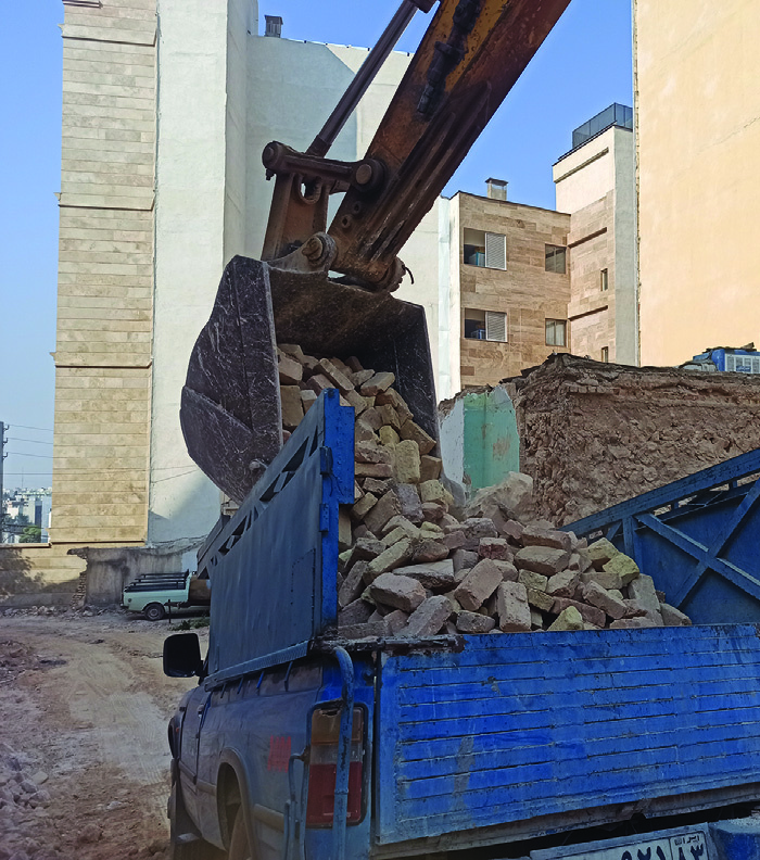 An excavator loading salvaged bricks from a demolished building onto a blue truck, the raw materials for the worker's house