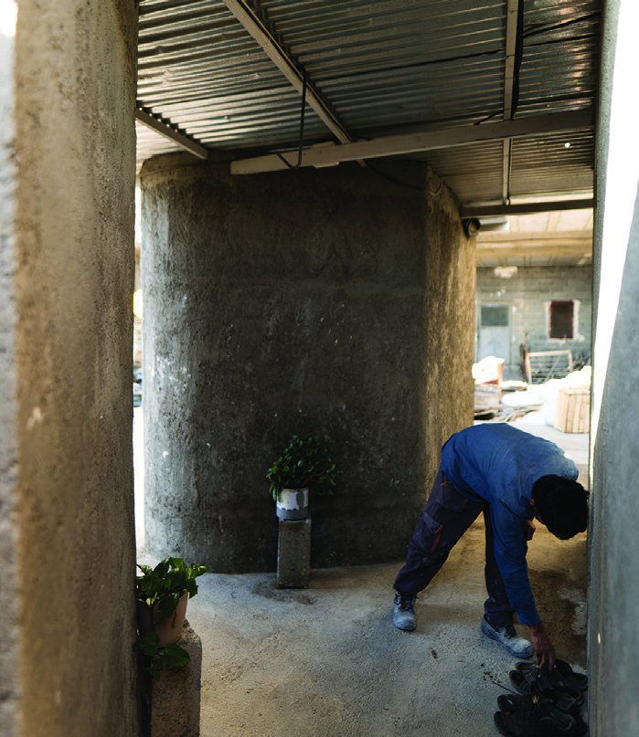A worker in the semi-open corridor between the curved concrete modules, with corrugated roof overhead providing shade, plants in the corridor
