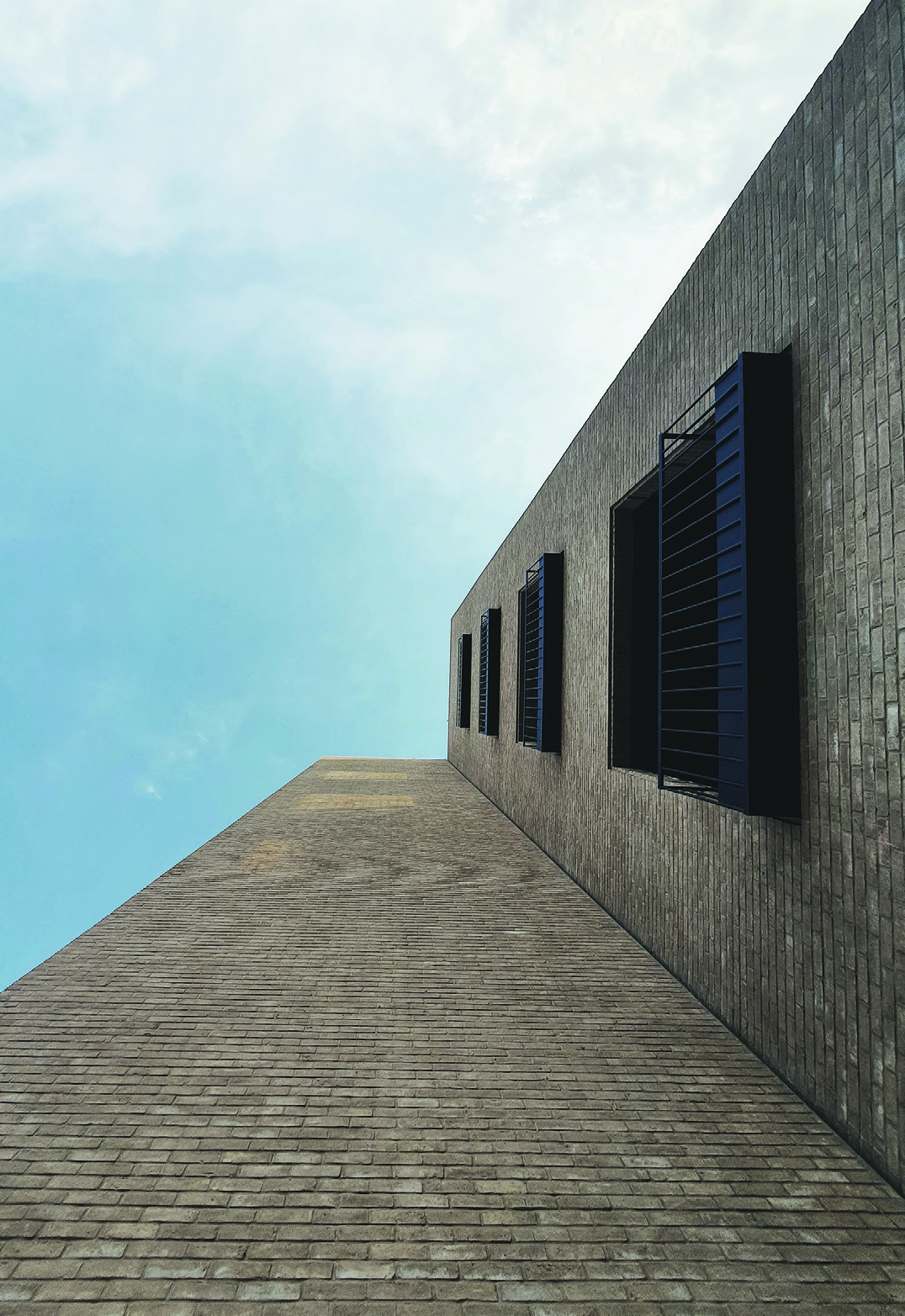 Low-angle view looking up along the brick side wall of the Sahra Building, showing dark metal window frames against the Kerman sky