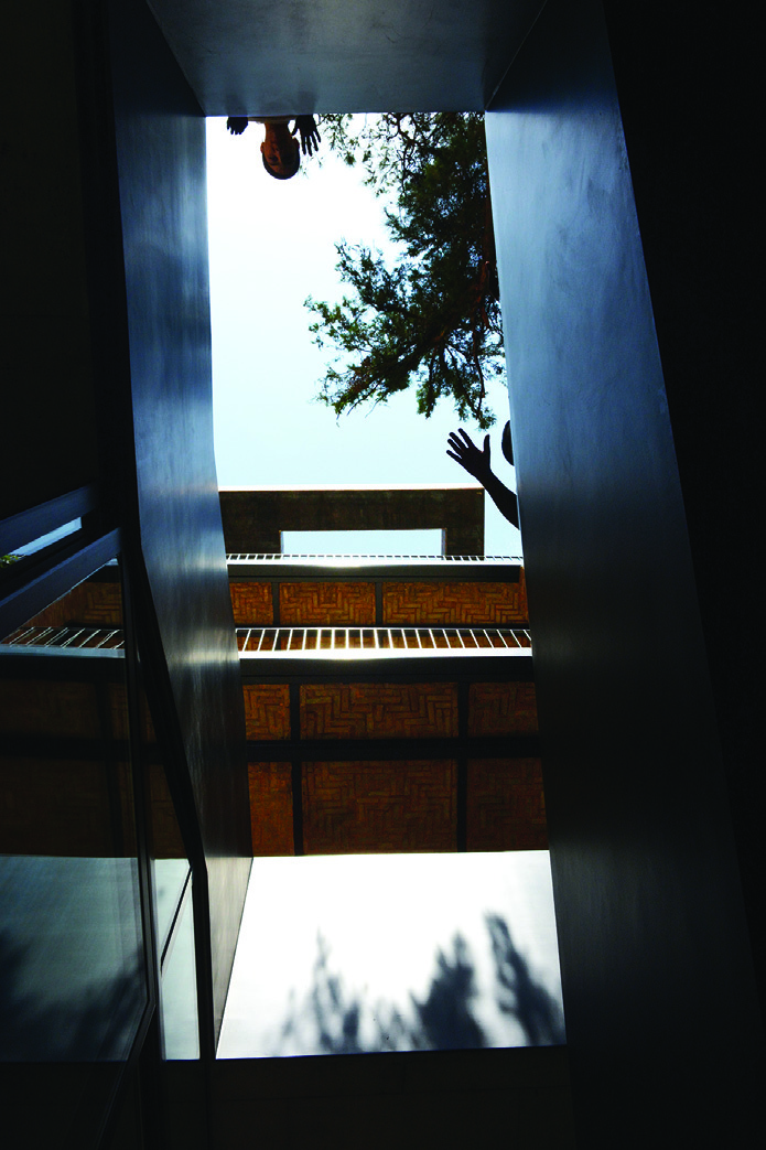 Vertical view up through the stairwell, showing concrete walls, herringbone brick detailing, a reaching hand silhouetted against the sky, and tree branches above