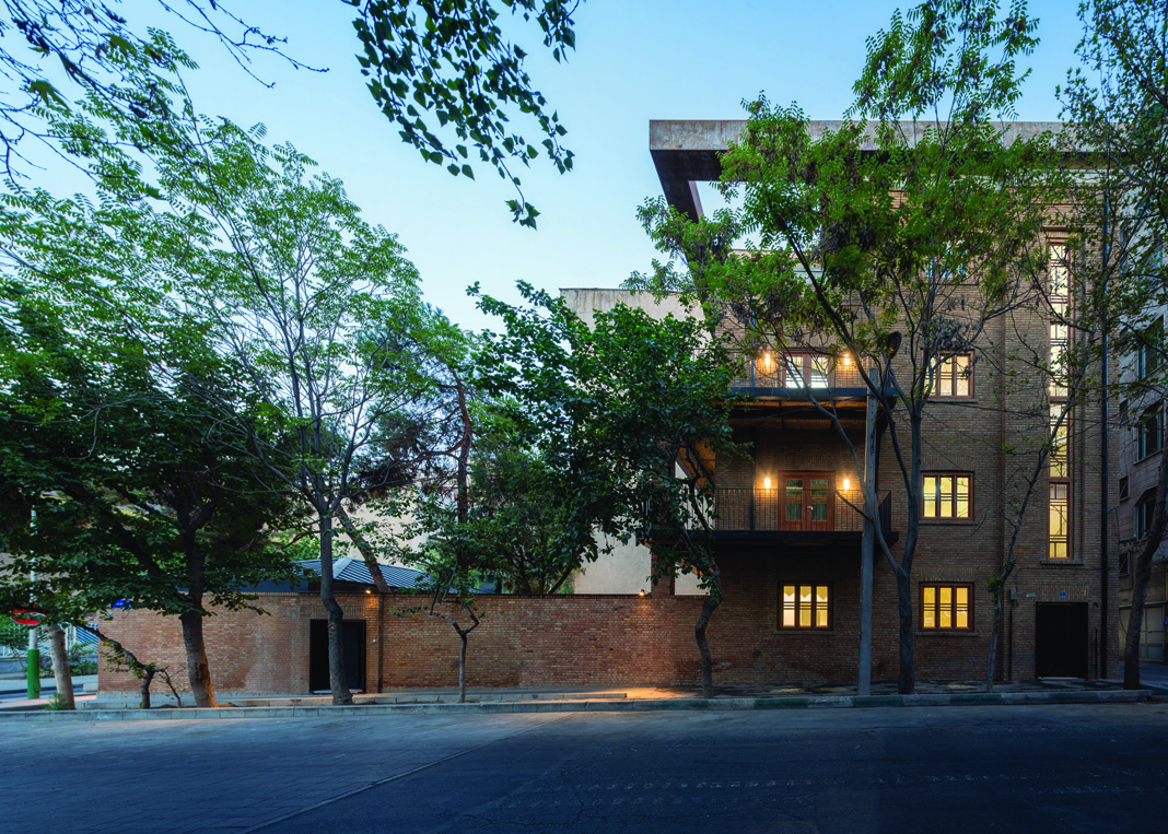 Street facade of the Khark Multipurpose Center at dusk, showing the brick building with concrete extension, illuminated interior, and mature trees lining the sidewalk