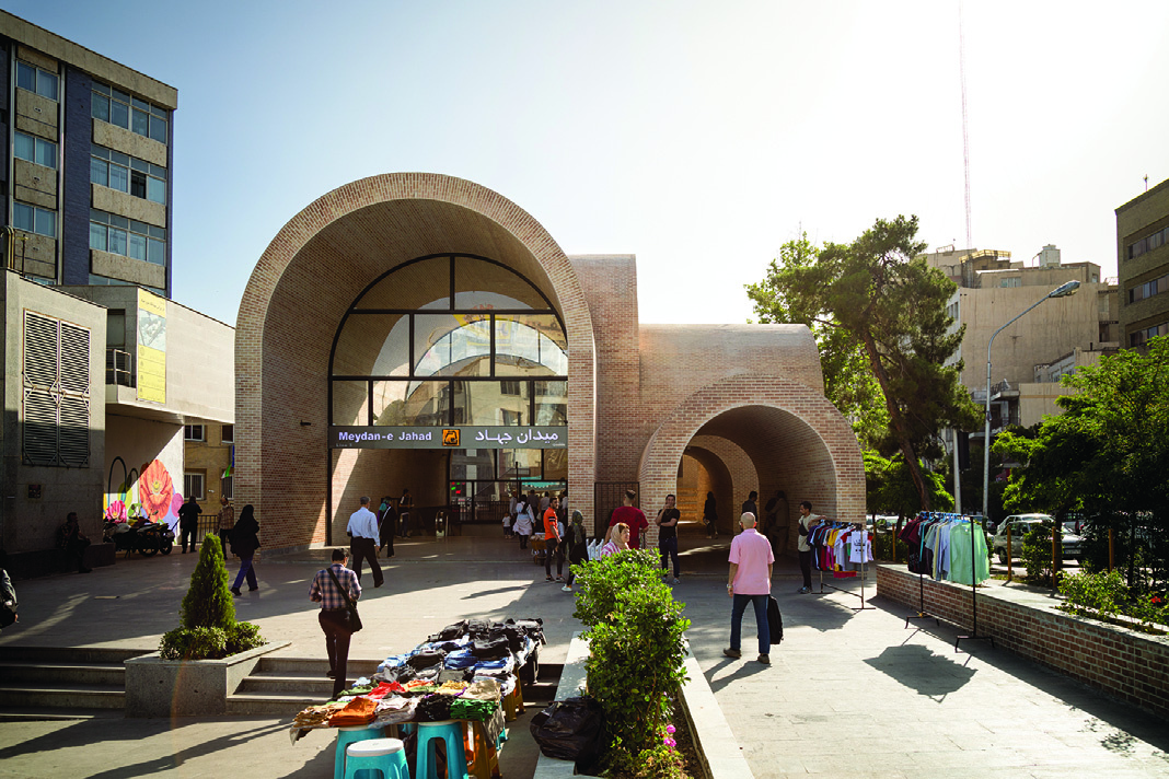 Street-level view of the metro entrance with the Meydan-e Jahad sign, brick arches, pedestrians entering and exiting, street vendors with goods displayed on tables, and green landscaping