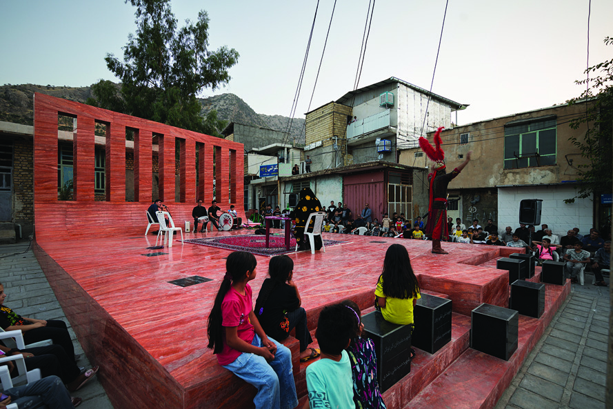 Ground-level view of a Taziyeh performance on the red platform at dusk, with children watching from the edge of the stage, musicians and performers on the platform, the red wall backdrop, and surrounding buildings visible