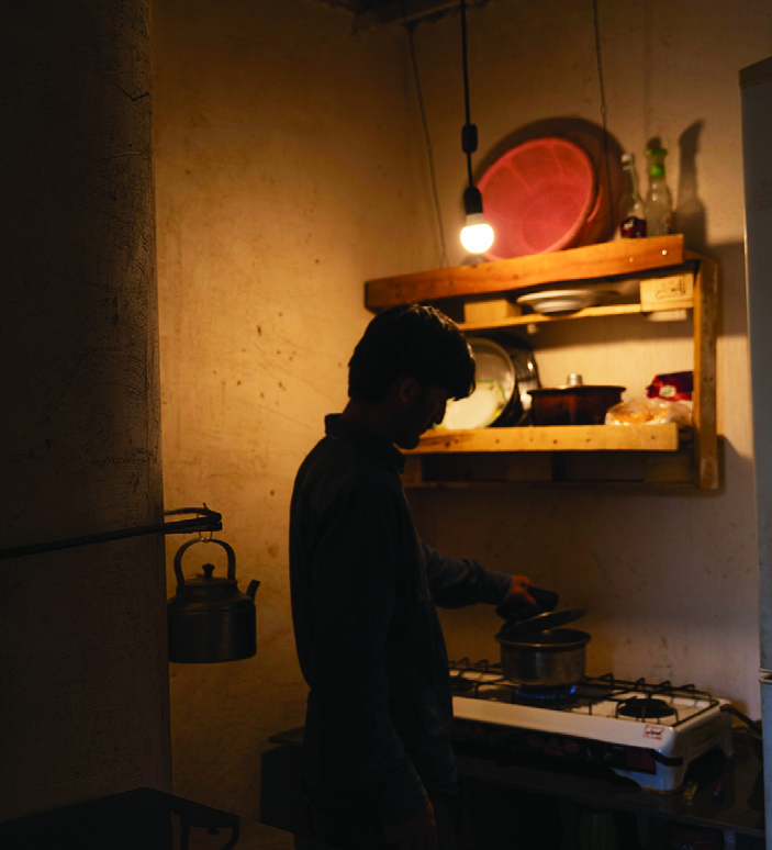 A worker cooking in the small kitchen corner with basic utensils hung on the wall and a single gas burner