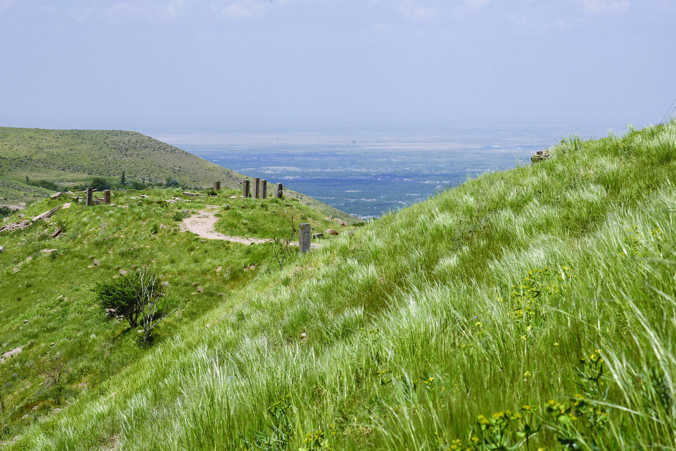 The ancient cemetery of Badamyar on a green hillside with standing gravestones, overlooking the vast Dehkharqan plain and Azarshahr in the distance