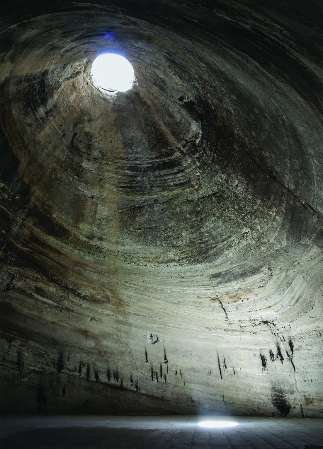 Looking up inside the Badamyar Mithraeum: the conical ceiling with layered geological strata and candle soot marks rises to a bright oculus at the apex, with light falling on the curved walls