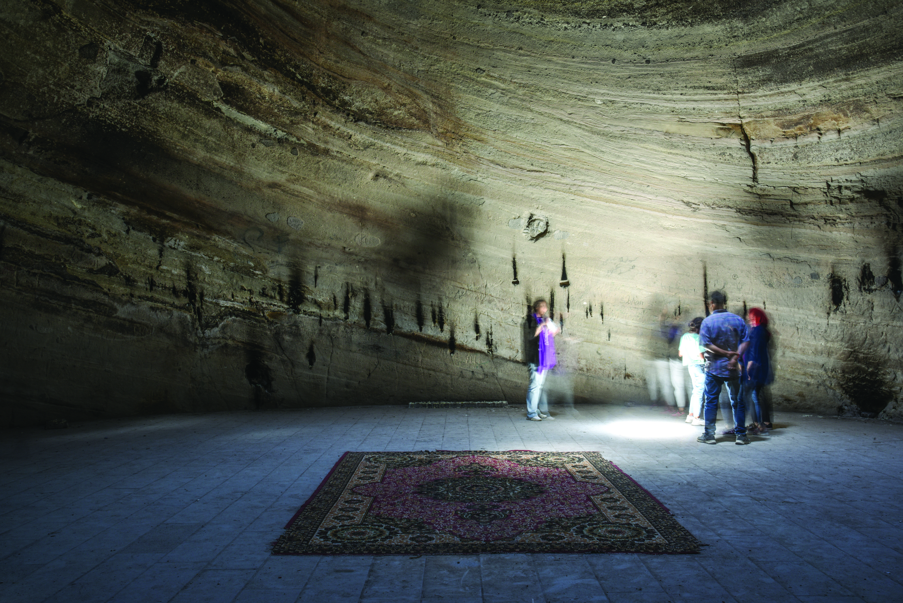 Visitors inside the Badamyar Mithraeum stand beneath the conical rock chamber with layered geological strata visible on the walls and candle soot marks; a carpet lies on the tiled floor
