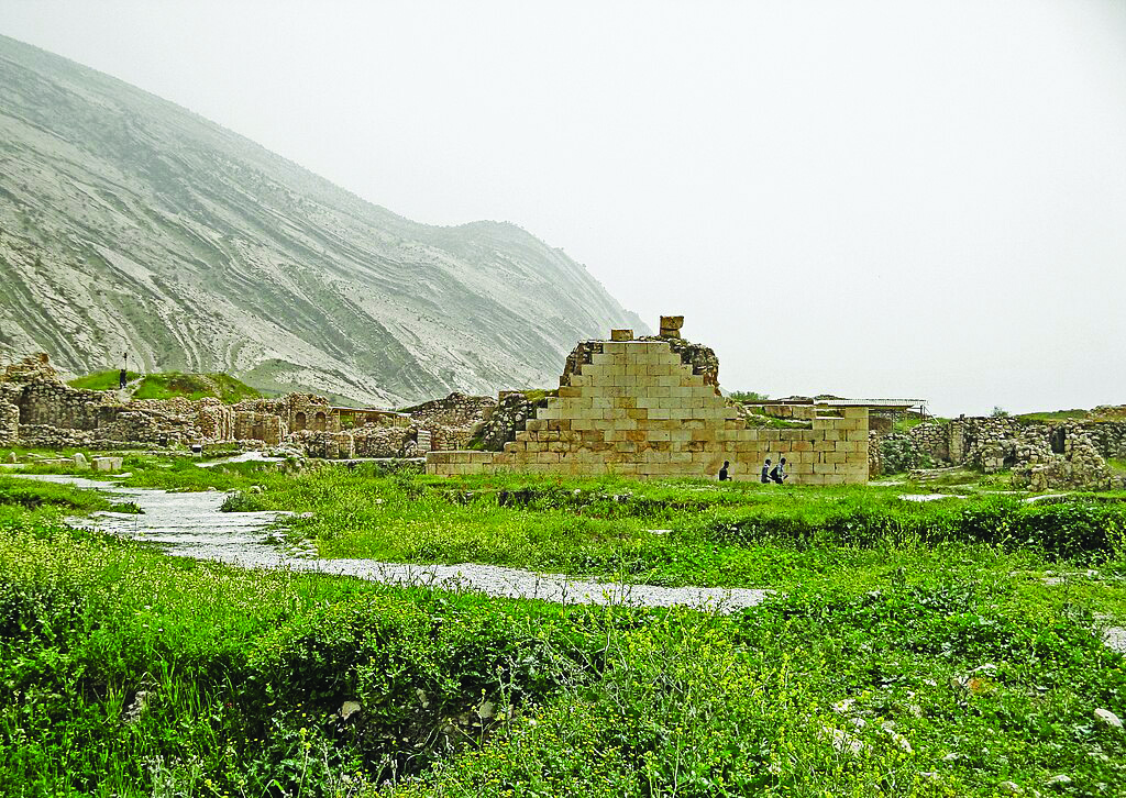Ruins of the Anahita temple at Bishapur with a dramatic mountain backdrop, showing the stepped stone structure amid green vegetation