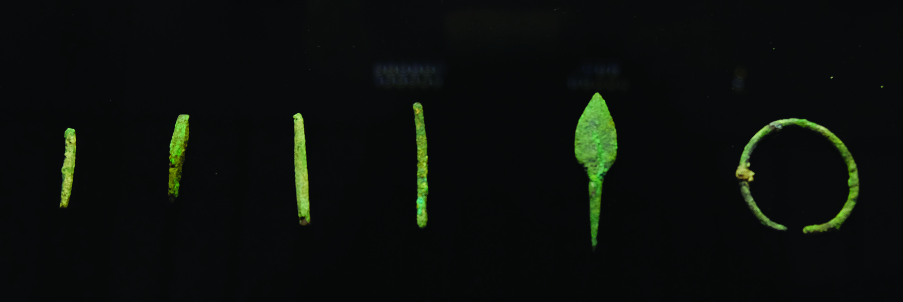 A row of ancient corroded bronze tools displayed against a black background, including needles, an arrowhead, and a bracelet, all showing green patina